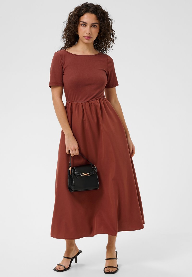 Femme aux cheveux bouclés portant une robe midi à manches courtes couleur rouille, tenant un petit sac à main noir, et portant des sandales à talons noires.