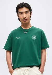 Νέος άνδρας που φοράει πράσινο μπλουζάκι με λευκό κείμενο και λογότυπα, μπεζ παντελόνια και μαύρο κολιέ με ασημένιες χάντρες.