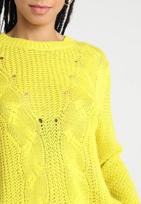 Jasnożółty sweter z dzianiny z wzorem teksturowym i detalami w otwartym splocie. Okrągły dekolt i długie rękawy. Miękki materiał, lekka konstrukcja.