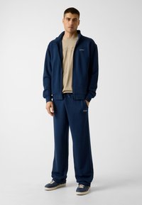 Marineblauer Sweatshirt mit Reißverschluss und Jogginghose aus weichem Material, mit "GUESS"-Logo auf Brust und Hose. Abgerundet durch legere Sneaker.