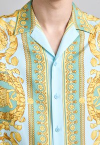 Short-sleeve light blue shirt featuring intricate yellow baroque patterns, with a pointed collar and blue buttons down the front.