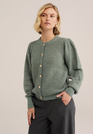 Blonde Frau mit kurzen Haaren, die goldene Creolen, eine grüne, geknöpfte Strickjacke mit Puffärmeln und dunkelgraue Hosen trägt, eine Hand in der Tasche.