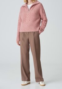 Femme portant un pull en maille torsadée rose à fermeture éclair, un pantalon large marron et des baskets blanches, debout avec une main dans la poche.