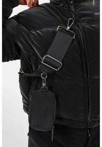 Zwarte nylon schoudertas bevestigd aan een zwarte riem, met metalen clips en een ritscompartiment met een textuurafwerking.