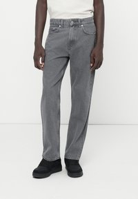 Jeans in denim grigio con taglio dritto, cinque tasche e cuciture sottili. Indossati con calzature nere, dotate di un colletto imbottito.