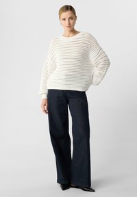 Weißer gestrickter Pullover mit lockerer Passform und weiten Ärmeln; kombiniert mit dunkle blauen, weiten Jeans; glattes, strukturiertes Material und offenes Gewebe-Design.