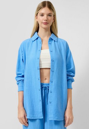 Donna bionda che indossa una camicia oversize azzurro chiaro abbottonata, pantaloni abbinati con coulisse, top corto in maglia bianco e orecchini a cerchio dorati.