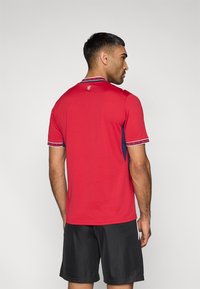 Κόκκινο αθλητικό πόλο μπλουζάκι με κοντά μανίκια, αντίθετες σκοτεινές μπλε πλευρικές πιέτες, μοτίβο για το γιακά και τα μανσέτα, και ένα μικρό λογότυπο στην πλάτη.