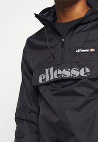 Mustan tuulenpitävän takin, jossa on puolivälin vetoketju, suuri harmaa "ellesse"-logo ja pieni värikäs logo rinnassa, valmistettu sileästä materiaalista.