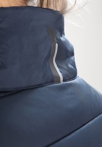 Mörkblå isolerad jacka med hög krage, med en slät yta och en reflekterande detalj längs sömmen.