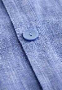 Šviesiai mėlynas lininis marškinių audinys su lygiu paviršiumi, turintis plastikinį mygtuką ir smulkius siuvimo detalių palei užsegimą.