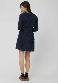 Robe longue bleu marine à manches longues avec broderie florale, taille cintrée, jupe à niveaux et poignets élastiques, assortie à des chaussures à plateforme marron et des chaussettes noires.