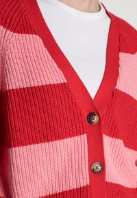 Randig cardigan i rött och rosa med djup V-ringning, ribbad textur och stora bruna knappar framtill. Bärs över en vit skjorta.