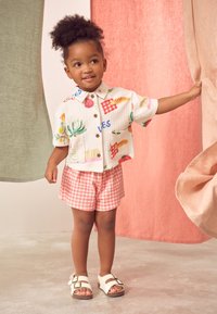Short-sleeve, collared shirt in cream with colorful fruit and vegetable patterns, paired with red gingham shorts and white sandals.