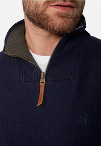 Marinblå stickad tröja med hög krage och dragkedja, med grå innerfoder och läderdragband, samt en subtil logodetalj på bröstet.