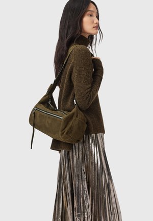 Olivgrüne Wildleder-Umhängetasche mit einer Reißverschlusstasche vorne, geschwungener Form und verstellbarem Riemen. Getragen mit einem dunklen Pullover und einem Metallic-Rock.