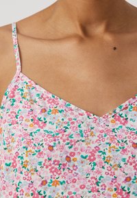 Detailní záběr horní části těla osoby, která má na sobě květinový top na tenké ramínka s růžovými, oranžovými, fialovými a zelenými květinami na bílém pozadí.