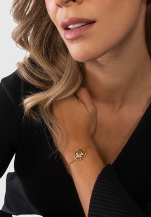 Frau mit welligem Haar, die ein schwarzes Oberteil trägt und ein goldenes Armband mit einem sonnenförmigen Design und einem zentralen Edelstein am Handgelenk präsentiert.
