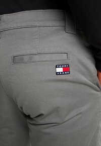 Šedé bavlněné kalhoty s kapsou na zadní straně, na které je modrá, červená a bílá nášivka "TOMMY JEANS". Hladká textura, přiléhavý střih a prošívané detaily.