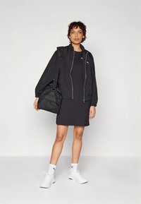 Giacca bomber leggera nera sopra un vestito nero, abbinata a sneakers bianche e una borsa a tracolla nera. Texture liscia e design elegante.