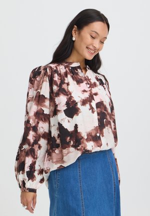 Femme souriante les yeux fermés, portant un chemisier rose et marron à motifs avec manches bouffantes et une jupe en jean bleue, sur un fond uni.