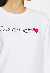 Felpa bianca con la scritta "Calvin Klein" stampata in nero, accentuata da un cuore rosso e un contorno a punti, realizzata in un materiale morbido e testurizzato.