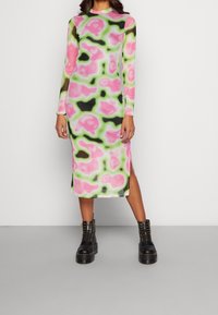 Robe à manches longues avec un motif abstrait rose, vert et noir, fabriquée en tissu doux avec des fentes sur les côtés. Associée à des bottes noires épaisses.