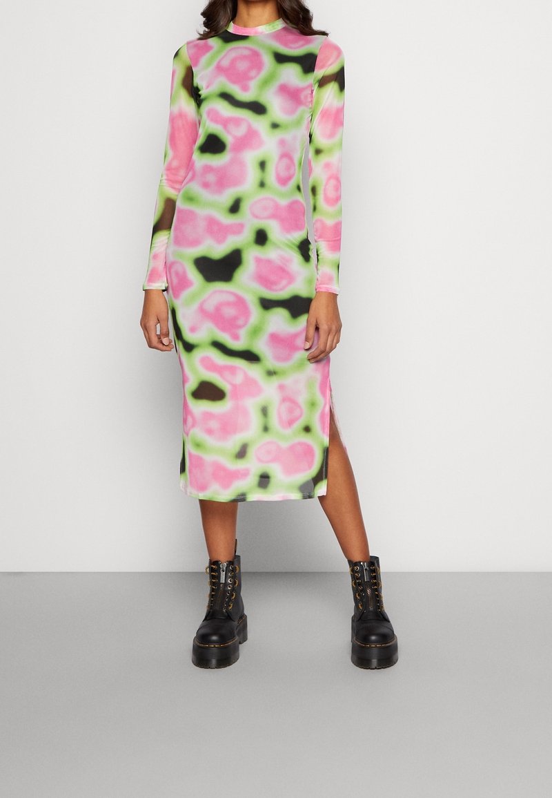 Robe à manches longues avec un motif abstrait rose, vert et noir, fabriquée en tissu doux avec des fentes sur les côtés. Associée à des bottes noires épaisses.