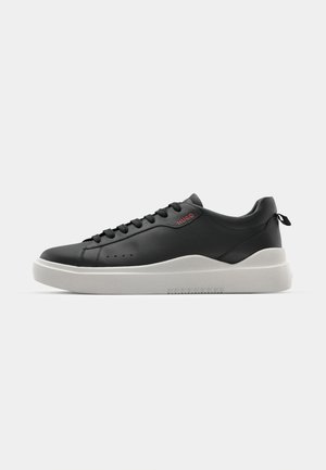 Sneaker nera in pelle liscia, caratterizzata da una suola grigia testurizzata, punta rotonda e lacci neri. Presente un piccolo logo rosso "HUGO".