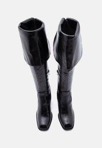 Paire de bottes hautes en cuir noir avec bouts carrés et larges revers pliés, présentées en vue de dessus sur un fond blanc.