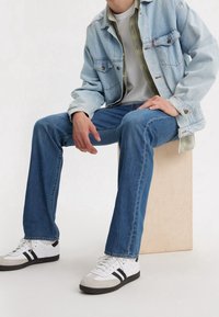 En ljusblå jeansjacka över en vit t-shirt, kombinerad med blå jeans och vita sneakers med svarta ränder. Sitter på en träkub.