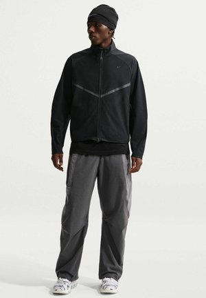 Mand iført sort jakke og hue, grå bukser og hvide sneakers, står mod en lys, ensfarvet baggrund og kigger til siden.