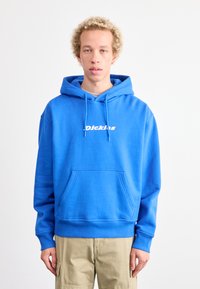 Sudadera de algodón azul con bolsillo canguro, puños acanalados y dobladillo. Presenta un logo blanco en el pecho. Combinada con pantalones cargo beige.