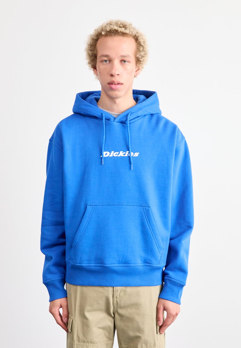 Sudadera de algodón azul con bolsillo canguro, puños acanalados y dobladillo. Presenta un logo blanco en el pecho. Combinada con pantalones cargo beige.