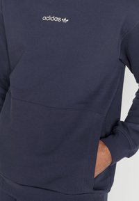 Person med en marinblå Adidas-tröja med vitt logo, handen i framfickan, visad från bröstet till höfterna.