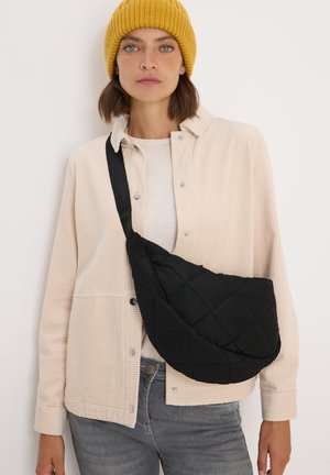 Black quilted sling bag with a textured surface, worn crossbody. The model wears a cream corduroy shirt and light gray jeans. Yellow knit beanie.