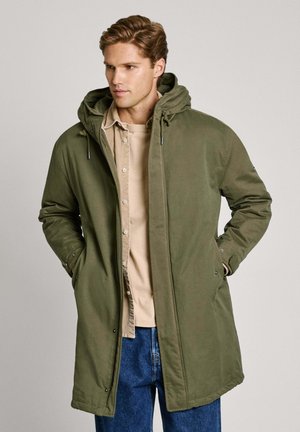 Olijfgroene parka jas met een capuchon, knopen aan de voorkant en zijzakken; gemaakt van duurzame stof; gecombineerd met een beige shirt en blauwe spijkerbroek.