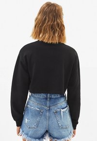 Mujer con cabello ondulado hasta los hombros, vistiendo un sudadera corta de manga larga negra y shorts de mezclilla desgastados de tiro alto.