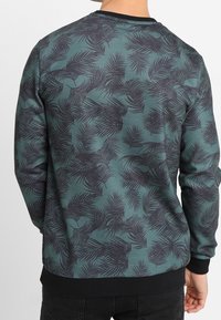 Felpa verde con motivo di foglie di palma scure. Presenta una scollatura tonda e polsini e orlo a coste neri. Tessuto morbido e vestibilità casual.