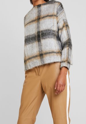 Donna che indossa un maglione a quadri peloso grigio, nero e beige con pantaloni beige caratterizzati da una striscia laterale bianca, su uno sfondo grigio semplice.