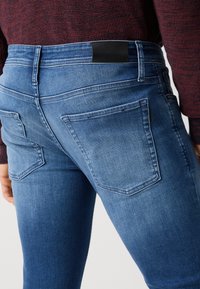 Jeans de mezclilla azul deslavado con cinco bolsillos, costuras visibles y un parche de cuero negro en la cinturilla. Primer plano de la vista trasera.