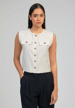 Femme debout, main dans la poche, portant un haut sans manches beige avec des boutons dorés et un pantalon bleu marine à rayures contre un fond gris.