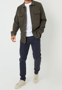 Tummanvihreä napillinen takki, jossa kaksi rintataskua, valkoinen t-paita alla, yhdistetty tummansinisiin housuihin ja vaaleanvärisiin lenkkareihin.