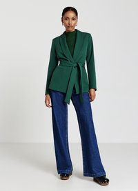Blazer verde con cintura, dotato di colletto a punta, abbinato a jeans in denim blu a gamba larga. Sotto, un top in maglia strutturato aggiunge profondità. Scarpe nere con platfond completano il look.