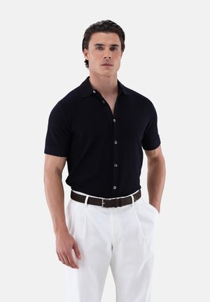 Mann trägt ein kurzärmeliges schwarzes Hemd mit Knöpfen, das in weiße plissierte Hosen mit einem dunkelbraunen Gürtel gesteckt ist, steht mit einer Hand in der Tasche.