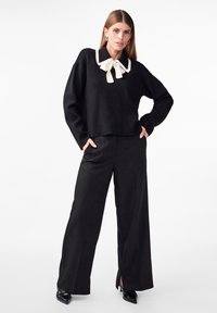 Pull court noir avec col blanc et nœud, associé à un pantalon large taille haute noir et des escarpins noirs à bout pointu.