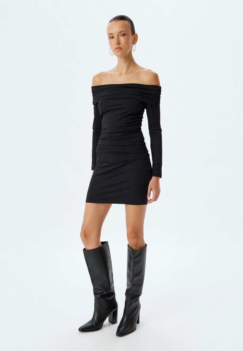 Schwarzes Off-Shoulder-Kleid mit langen Ärmeln und Raffung. Kombiniert mit knielangen schwarzen Lederstiefeln mit Blockabsatz.