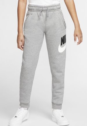 Persoon draagt grijze Nike joggingbroek met zwart-wit logo op dij, witte sneakers en een wit overhemd ingestopt, staand op een grijze achtergrond.