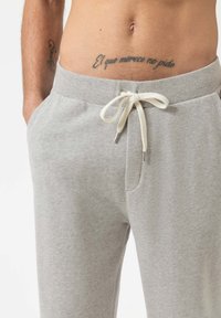 Blote torso met een Spaanse tatoeage boven grijze sweatpants met een witte trekkoord, handen in de zakken, en een kleine tatoeage op de linkeronderarm.