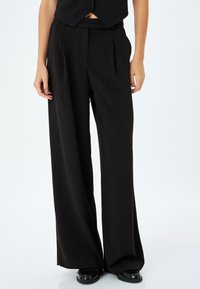 Zwarte wijde broek van gladde stof, met voorplooien en een hoge taille. Gecombineerd met een cropped zwart vest en platte schoenen.